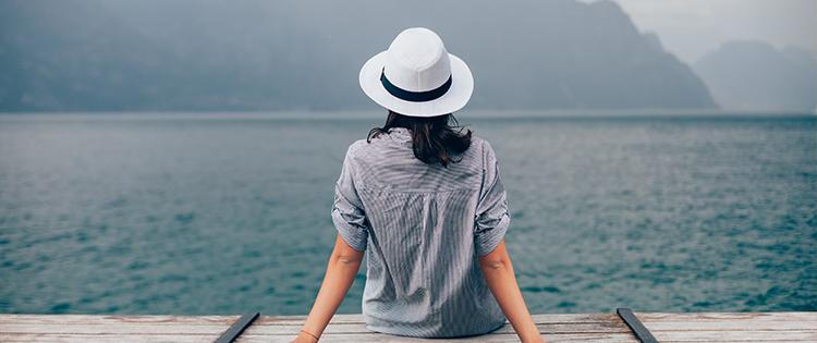 Photo of a woman wearing a hat while sitting on a pier and looking at the water.