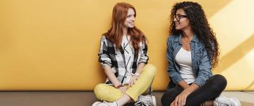 Photo of two young women sitting against a wall and talking.