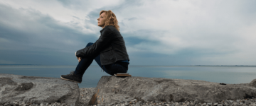 Photo of a blonde woman sitting on a rocky beach on a cloudy day.