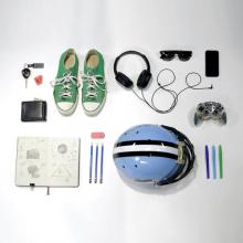 Lots of everyday objects are laid out on a white background. A wallet, car keys, a notebook, shoes, headphones sunglasses, pens and pencils, and even a video game controller are all set down in an orderly fashion. 